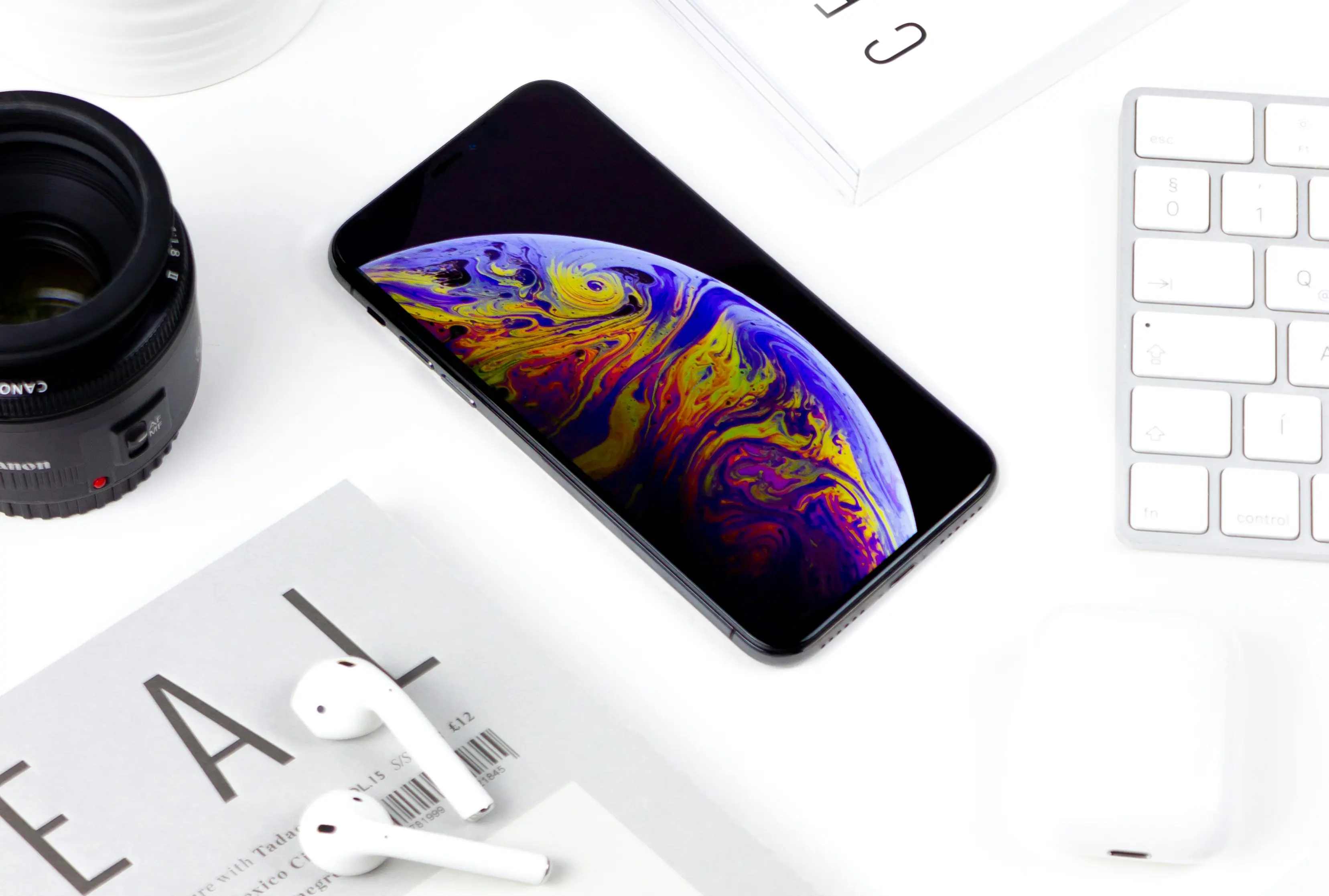 Presentation image featuring an iPhone with a colorful screen-saver, a bit of an Apple keyboard, a pair of AirPods placed on a magazine and a camera objective, all on a white background.