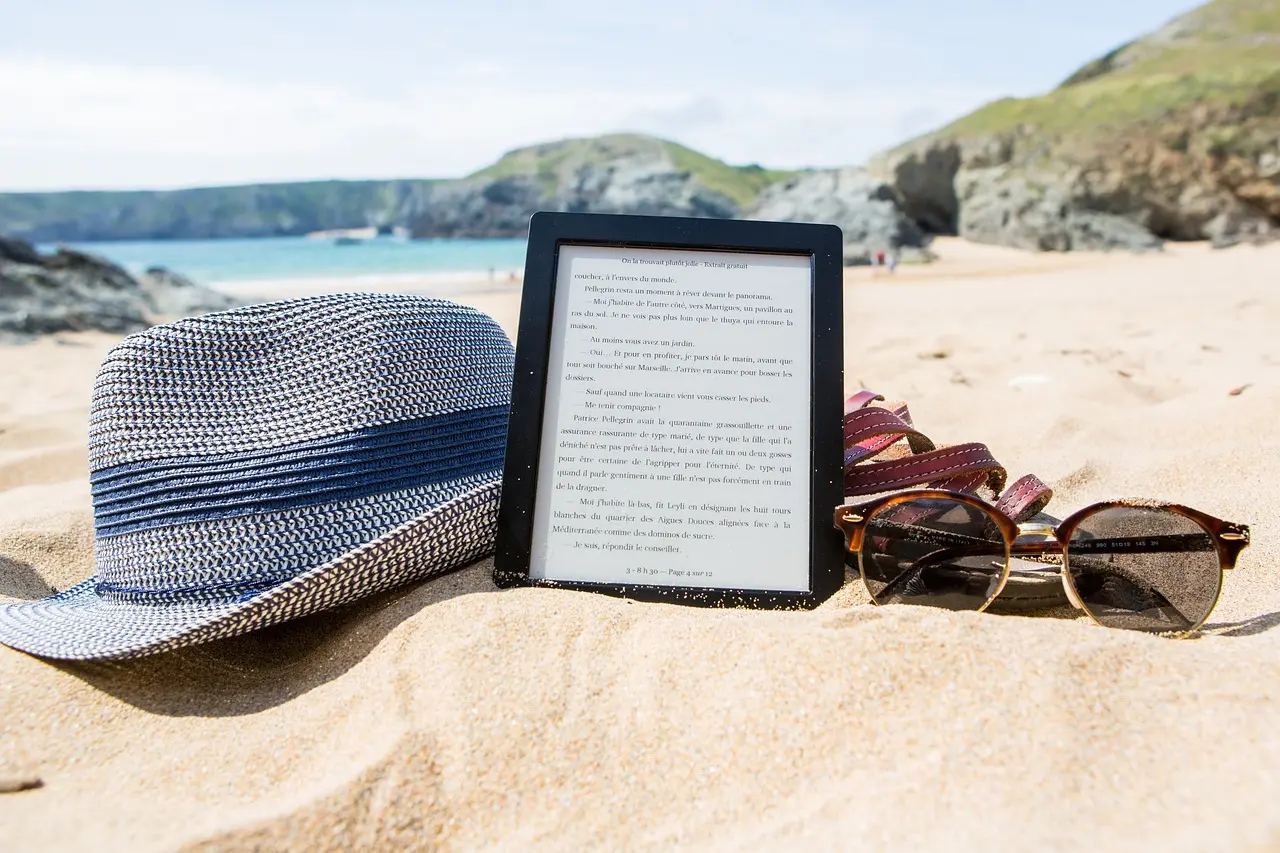 Presentation image featuring a sun hat, an open ebook reader, sandals and sunglasses on a sandy shore, with rocky hills and water in the background.