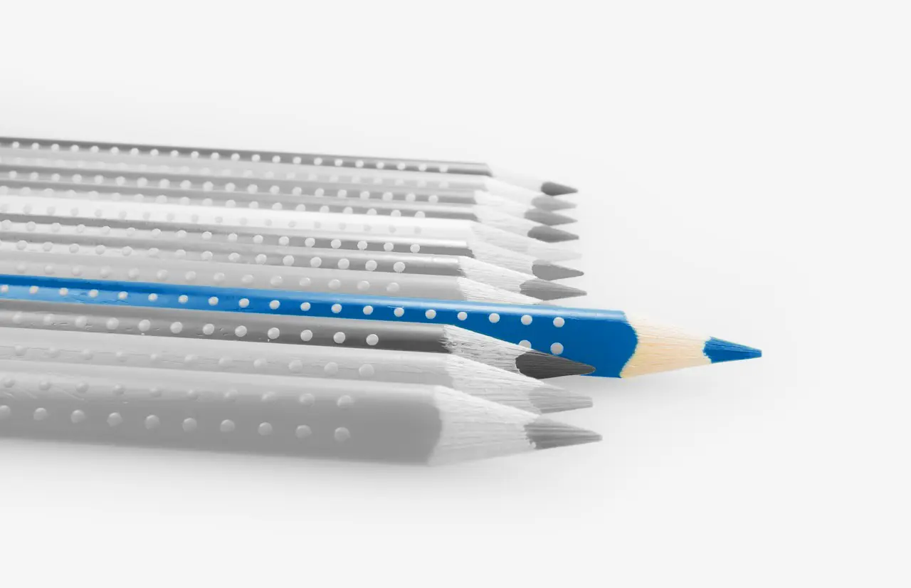 Black and white presentation image of a row of sharpened pencils neatly lined-up together, with a blue sharpened pencil standing out of the black and white row.